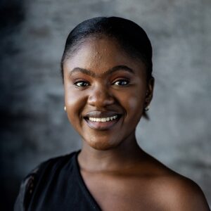 Portrait of Sisikelelwe Mngenela, Judith Neilson Young Artist at Cape Town Opera, wearing a black dress, smiling with her hair tied back, against a grey backdrop.