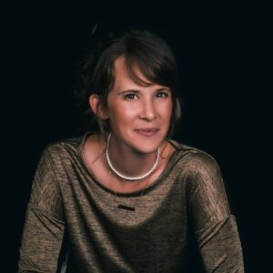 Professional headshot of Movement Director Cleo Notcutt-Williams. She has long dark hair tied up and is wearing a gold-toned blouse, a white necklace, and a gold necklace, smiling warmly into the camera.
