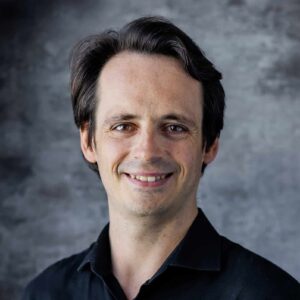 Profile portrait of Conductor Jan Hugo smiling with ear-length black hair.