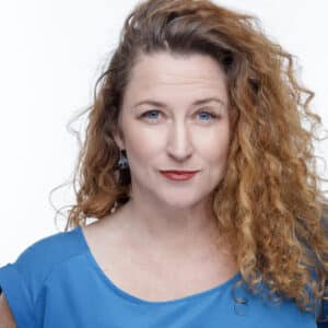 Professional studio headshot of Choreographer and Movement Director Fiona du Plooy. She has long, curly, blonde-reddish hair, bright blue eyes, bold red lipstick, and is wearing a light blue blouse while looking toward the camera.