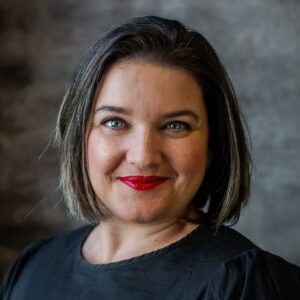 Professional studio headshot of Maritha Visagie, Head of Production and Costume Designer for Cape Town Opera. She has short hair styled above her shoulders, bold red lipstick, and is wearing a classic black blouse.