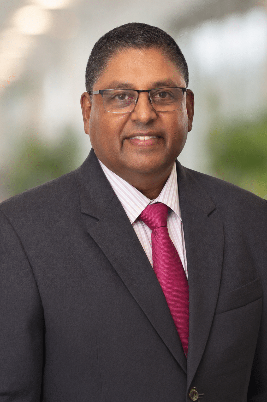 Professional studio headshot of Naushad Khan, wearing glasses, a grey formal blazer, a lightly pink striped dress shirt, and a red tie. He has short, neatly styled hair.